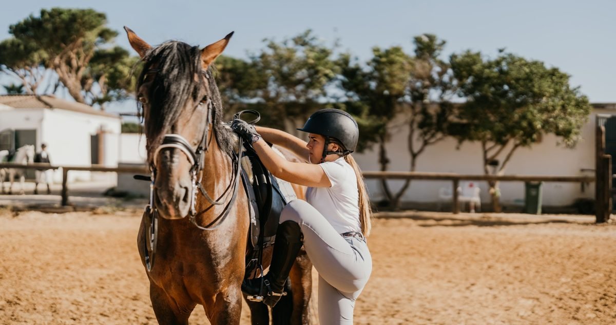 Young rider in a helmet mounting her horse in a sand arena, a moment that asks for the hip and knee control equestrian fitness builds.