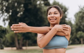 Woman easing into a cross-body shoulder stretch outdoors, the warm-up that keeps an irritated bursa from flaring.