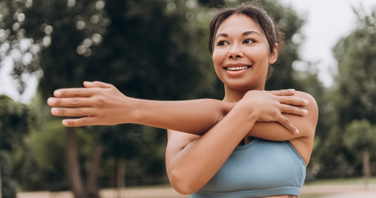 Shoulder Bursitis: Symptoms and When to See a Specialist Woman easing into a cross-body shoulder stretch outdoors, the warm-up that keeps an irritated bursa from flaring.
