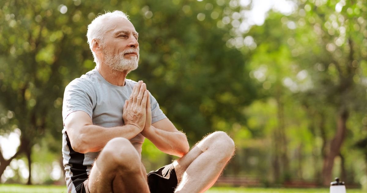 Staying active and mobile as you age Older man sitting cross-legged on the grass in a sunlit park with his hands together, staying active and mobile as he ages.